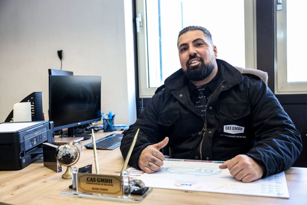 Mann im Büro, lächelt, trägt dunkle Jacke, Bart; Schreibtisch mit Globus und Schildern.